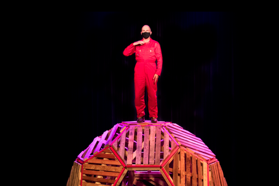 A person in a red jumpsuit stands on top of a wooden spherical structure