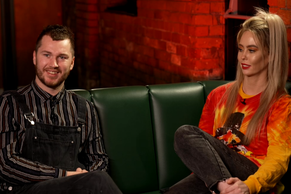 NoGood Boyo (2 band members) sat on a green couch with brick illuminated in red in the background.