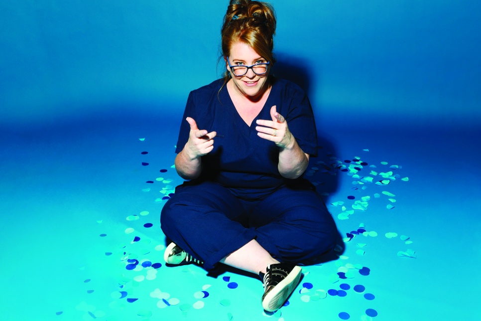 Comedian Georgie Carroll wears blue scrubs and point at the camera