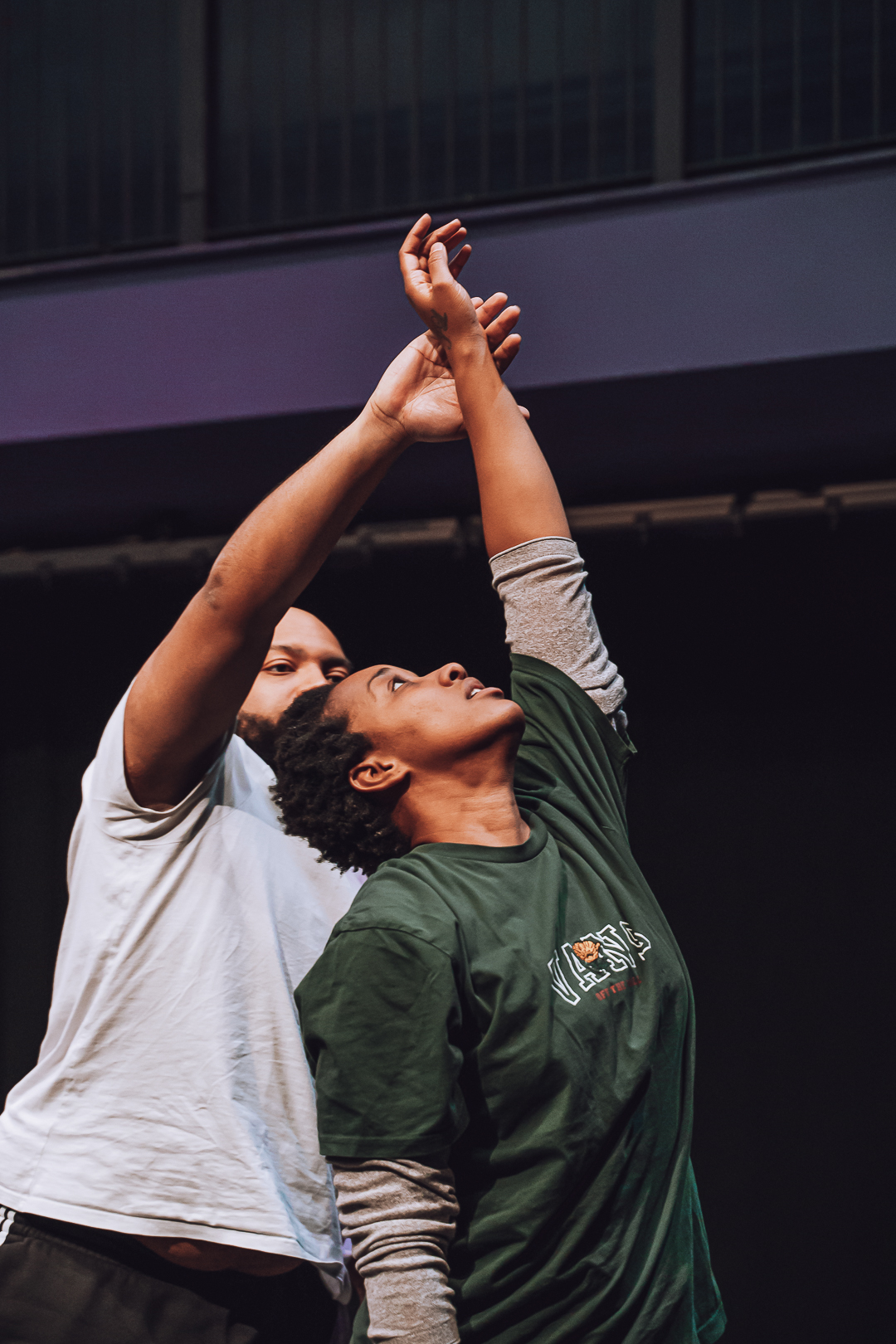  Two brown-skinned dancers standing back to front with their arms extending in the air gently touching. 