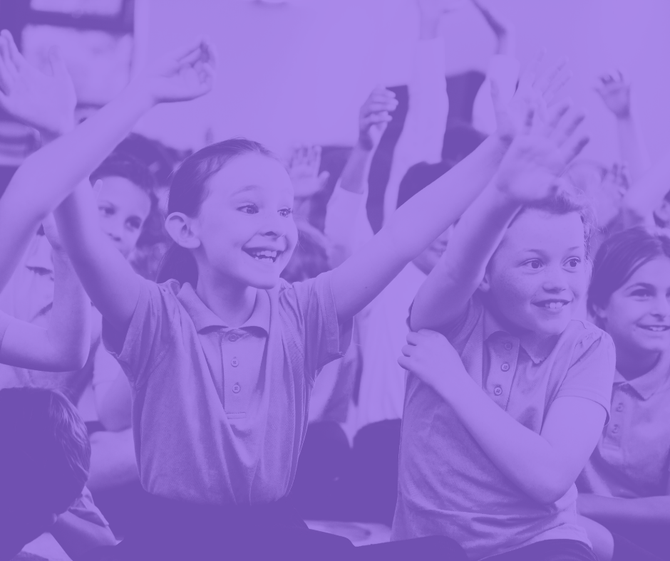 The image is of around 8 - 10 school children, they are sat down. There are 2 prominent/ in focus children who both look happy an engaged in something with their hands in the air. The whole image has a purple wash over it. 