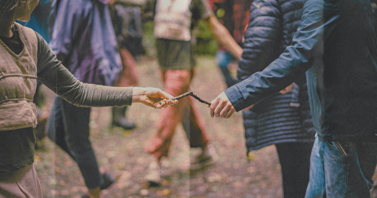 Image of people outdoors. There are two people in the foreground who are holding each end of the same wooden stick / Delwedd o bobl tu allan. Mae dau berson yn y blaendir yn dal yr un brigyn pren ar bob pen