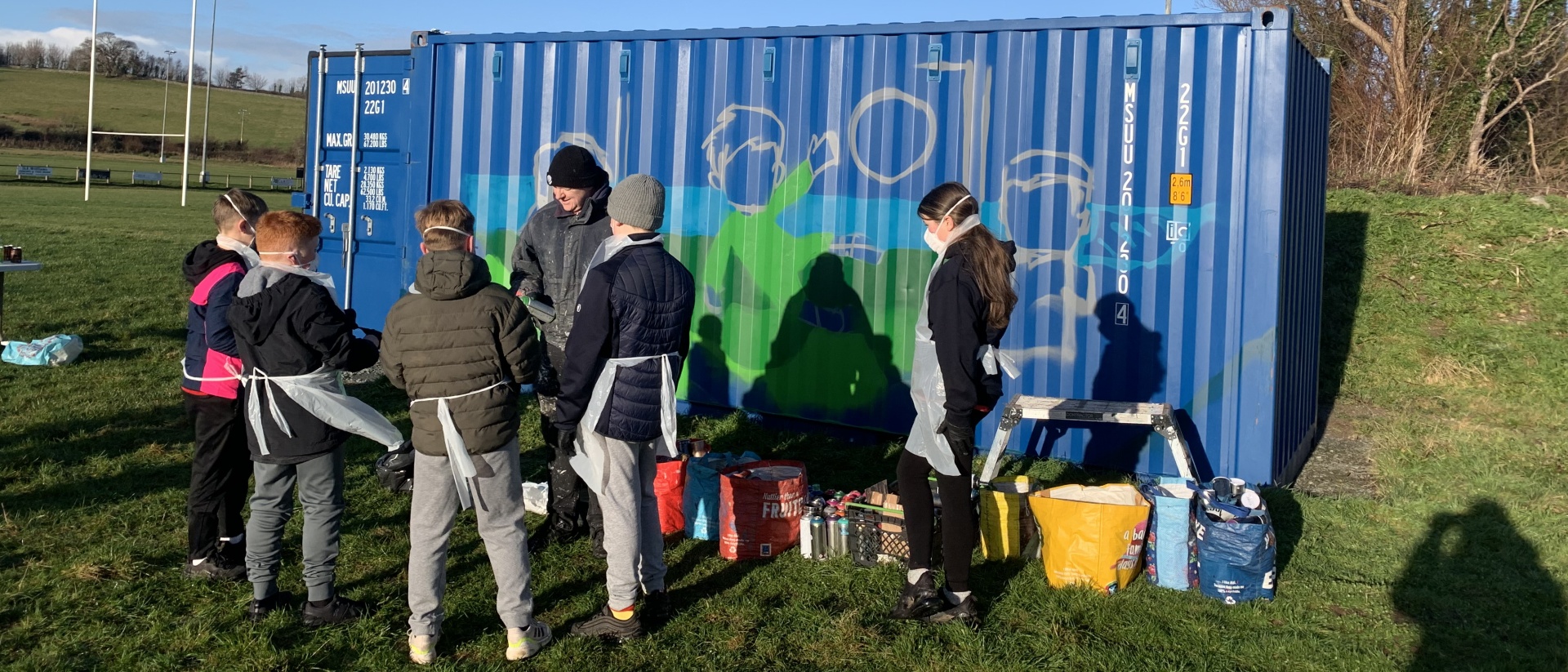 Young people and children painting a mural on a blue shipping container in a field