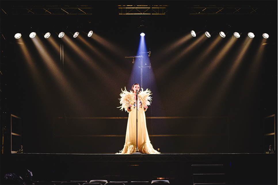 Lady stood centre stage in a spotlight, dressed in long, white, feathered gown