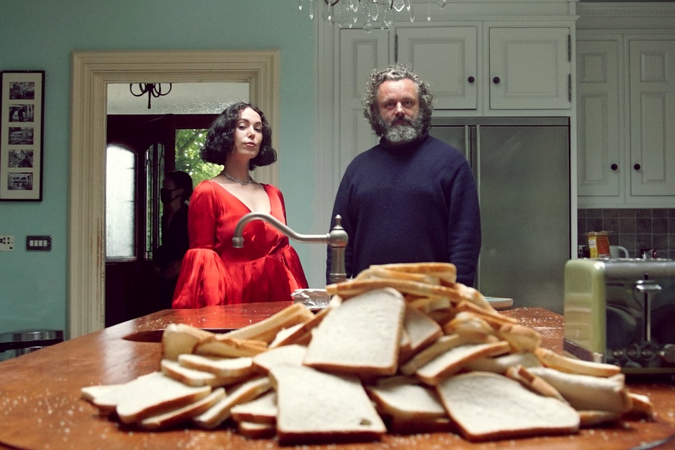 Kelly Lee Owens & Michael Sheen stood in a kitchen behind a large pile of bread