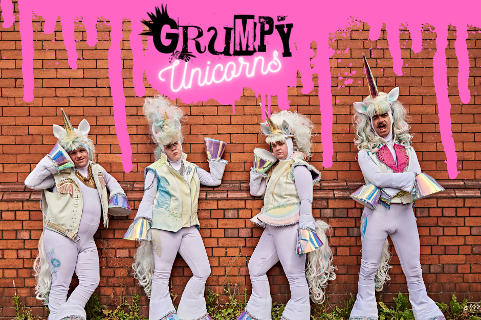4 people dressed as unicorns pose against a red brick wall with pink paint dripping down and 'Grumpy Unicorns' written above