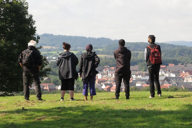 5 people stood in a field facing away from the camera overlooking a view of houses below them