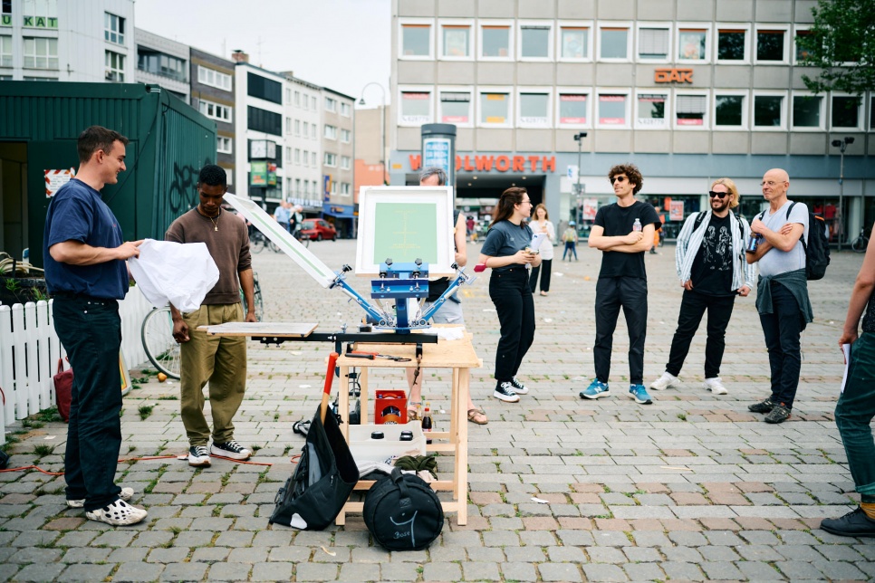 Group of young people stood around a screen printing set up in a city square