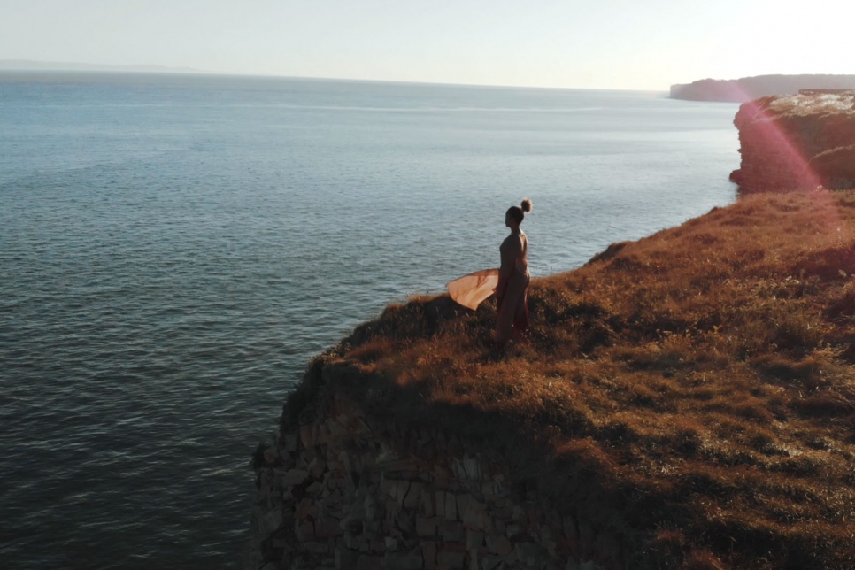 A performer stands at a grassy cliff edge overlooking the sea. They're stood facing the water, wearing a beige dress which is flowing in the wind and their hair is tied up.