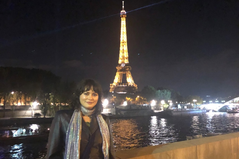 A person stood on a bridge over the river Seine at night-time with the Eiffel Tower lit up in gold behind them and white lights from the other buildings in the background