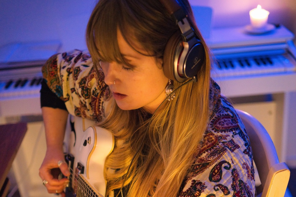 A person wearing headphones sat in a white chair playing a white guitar. Behind them is a white keyboard piano and on top of the piano is a lit candle