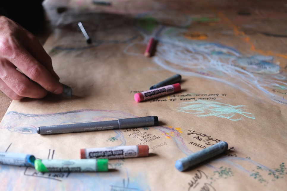 A hand is using a piece of chalk to draw on a brown piece of paper. There are many pieces of chalk in different colours laying on the brown paper.