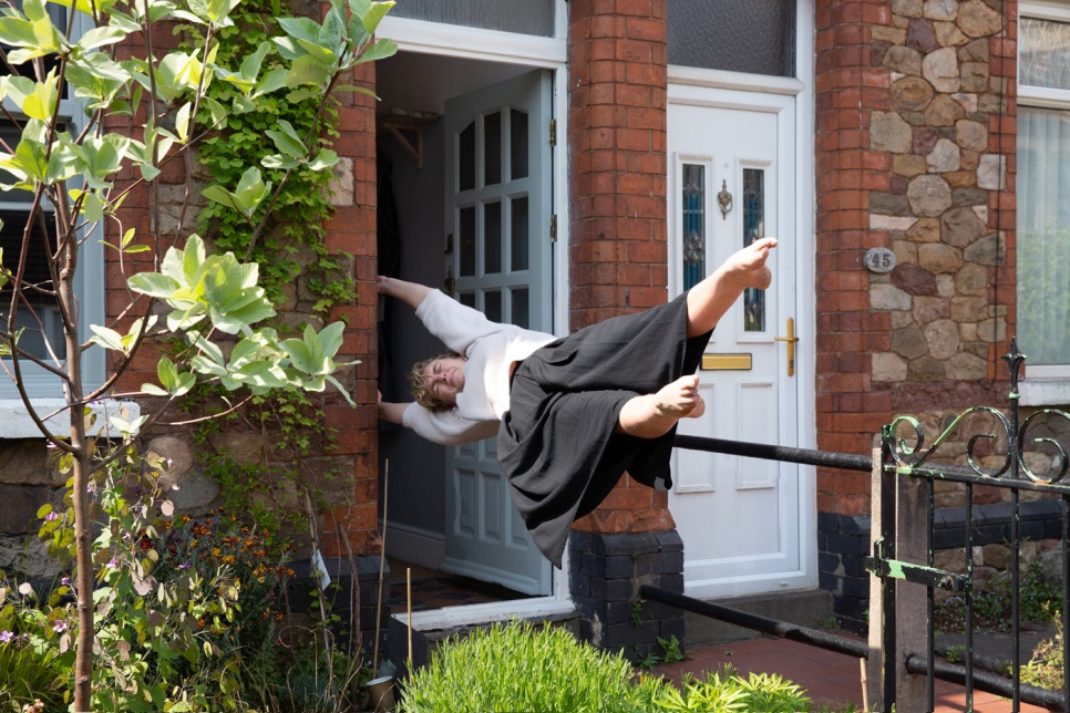 A person is gripping onto an open doorframe of a house as they float horizontally in mid-air. Their toes are pointed, and their face is grimaced, as if an invisible person is trying to drag them away from the house.
