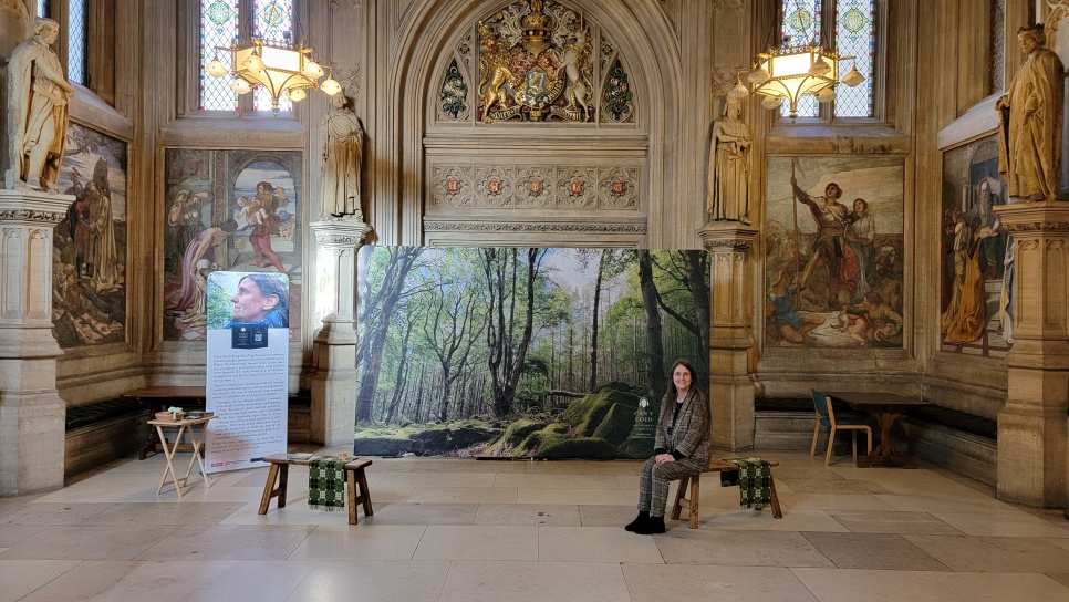 Lady sat smiling on a bench in front of a large exhibition photograph in a grand room, at the Houses of Parliament