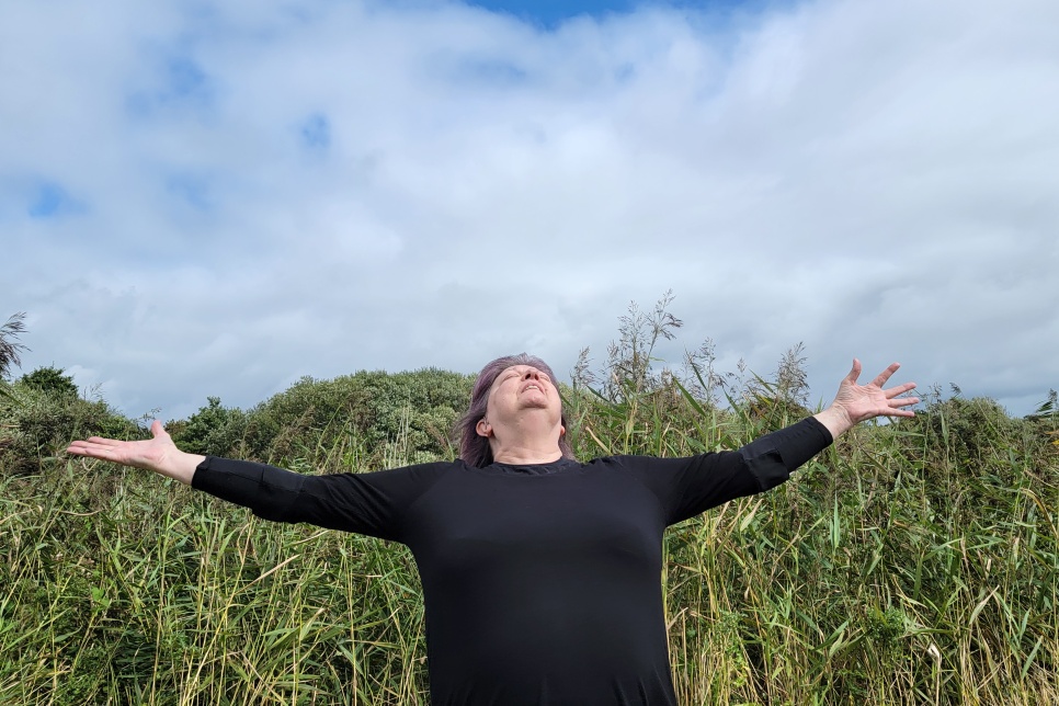 Lady in black top with purple hair, arms outstretched and eyes closed. Out in nature with green fields and blue sky.