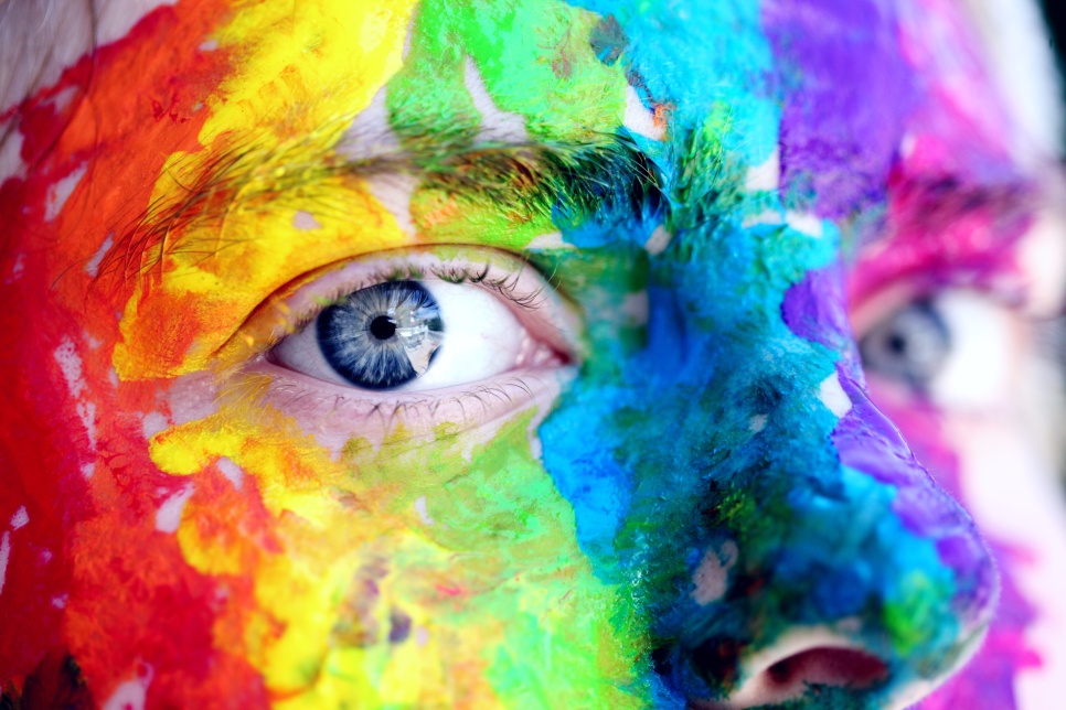A section of someone's face covered in multi-coloured paint looks to camera. 