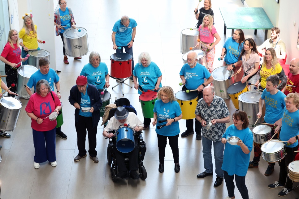 A group of people in brightly coloured T-shirts assembled in a large reception area playing Samba drums