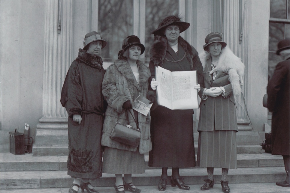 Black and white photo of 4 women in 1924 with hats and winter jackets, holding a large open book.