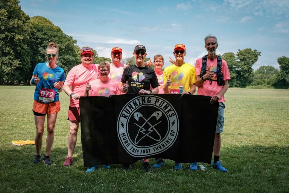 Running Punks - photo Anita Rice A group of people smiling in brightly coloured running clothing, on a sunny day in a field, holding a black 'Running Punks' flag.