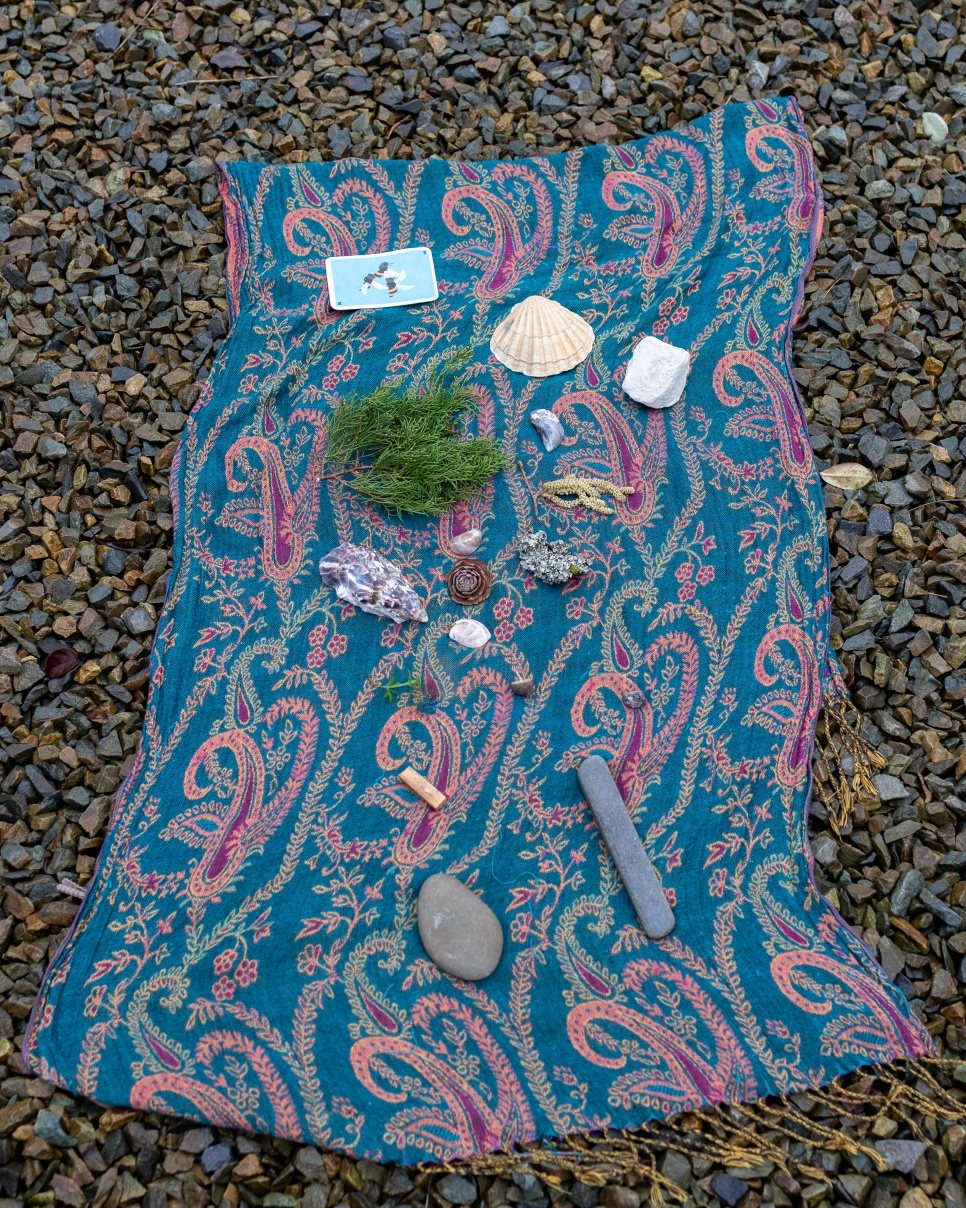 A Paisley patterned shawl on a pebble beach. On the shawl there is a playing card, a cigarette stump, some shells, pebbles and a fern