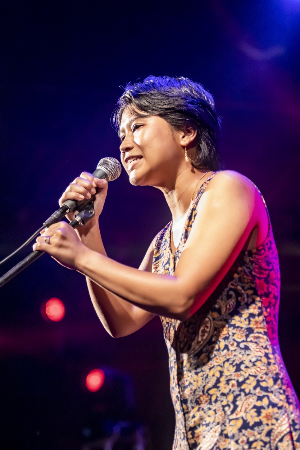 Lapdiang Syiem singing on a stage in a floral dress with a hand-held microphone on a stand 