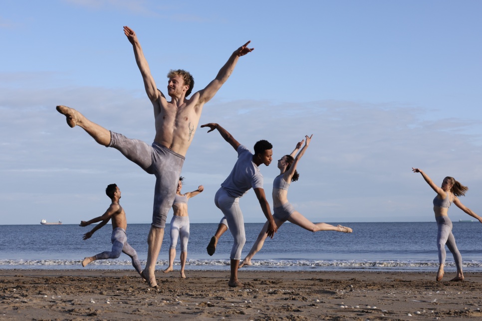 A group of Ballet Cymru dancers performing on a beach, showing dynamic poses against a backdrop of sea and sky.