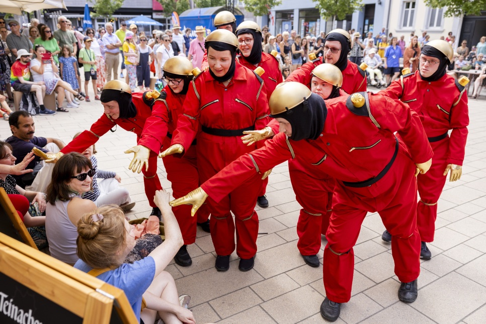 Actors performing as robots in an outside space dressed in red boilersuits and engaging with the audience