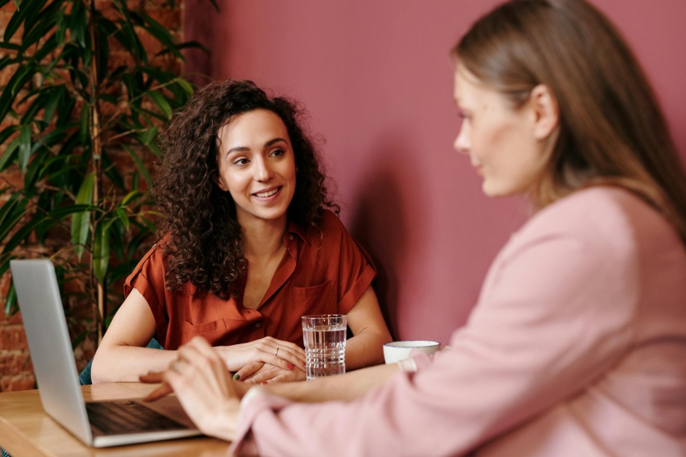 Two women sat at a small table working together on a laptop. The woman on the left is smiling and the woman on the right is typing on the laptiop.