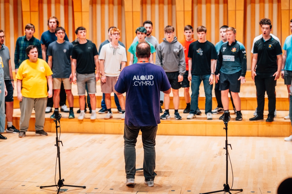 A man with a purple t-shirt that says ALOUD CYMRU on the back, is stood in front of a large group of a choir of boys in a wooden hall