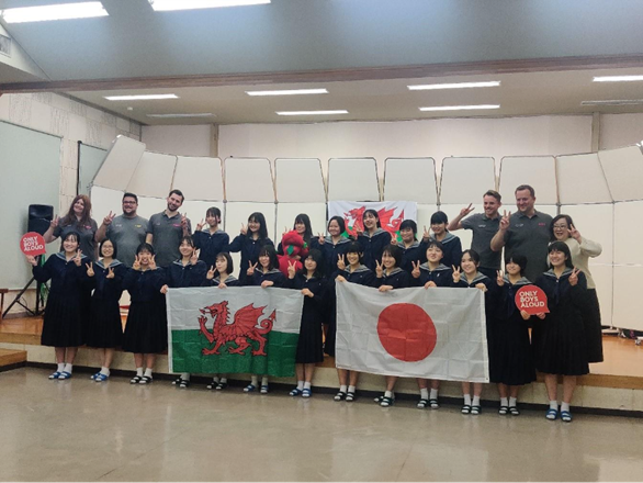 A group of schoolchildren in Japan and adults smiling and holding large Wales and Japan flags