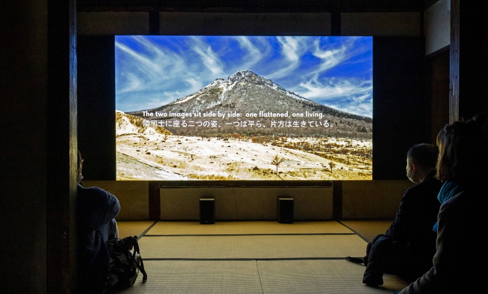 A still of a Japanese mountain with white writing over it, projected in a dark room in front of a seated audience.