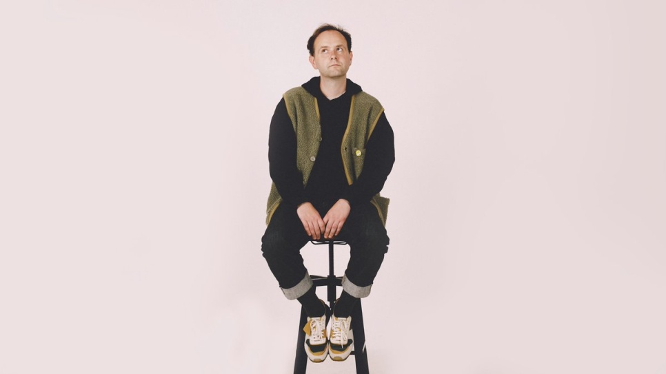 Man in olive green and black outfit, sat on a stool, looking upward, in front of a white background.