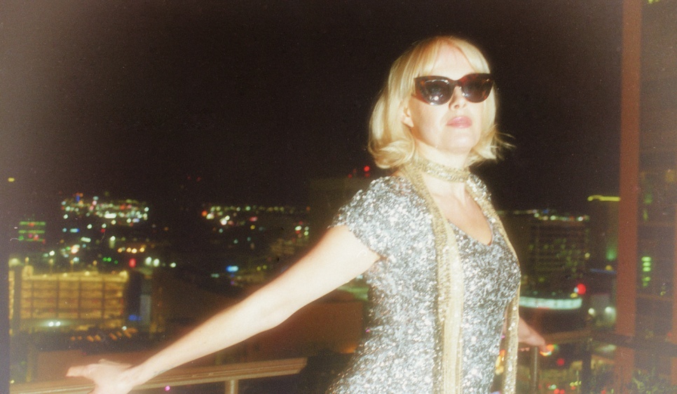 A woman with bobbed blonde hair and sunglasses in a silver sparkly dress in front of a night cityscape