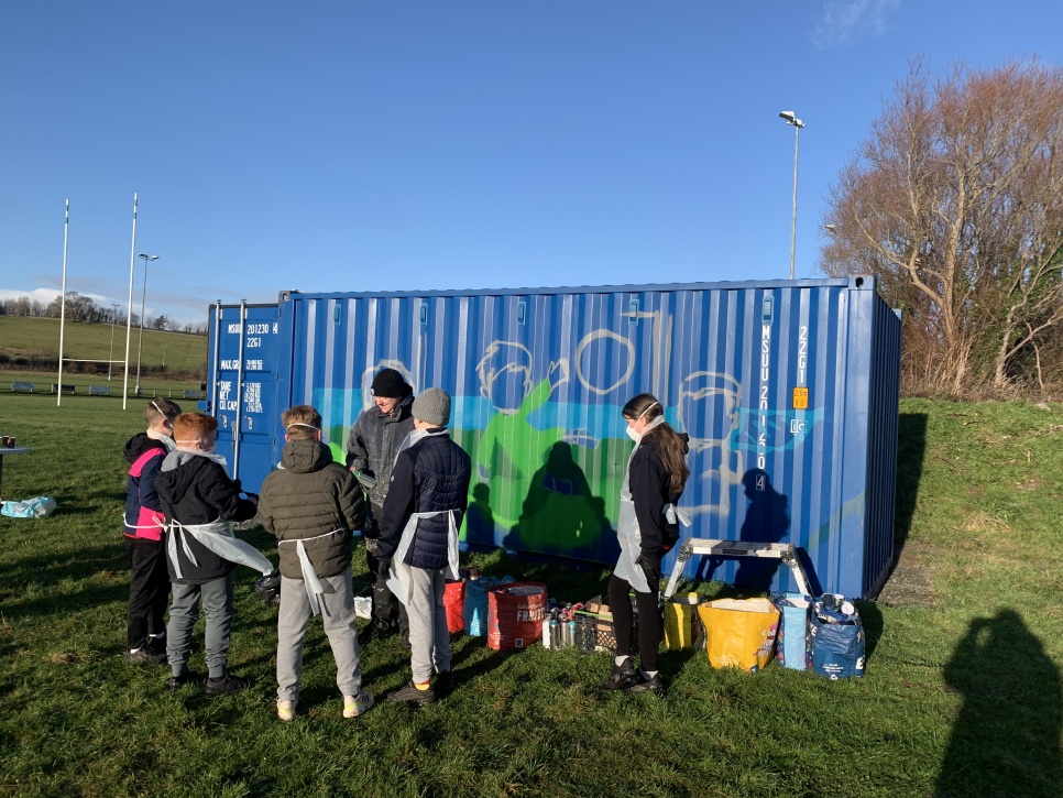 Young people and children painting a mural on a blue shipping container in a field