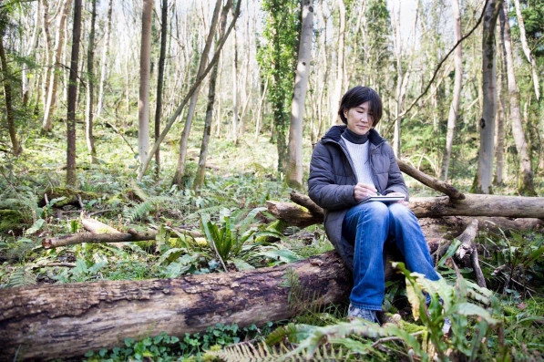 A woman with black bobbed hair sat on a fallen tree in a forest, sketching
