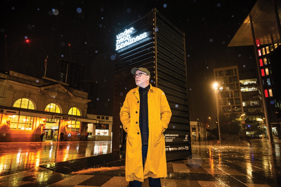 A man in a cap, glasses and yellow overcoat in a rainy city square at night with neon lighting that says 'Under Neon Lonliness'