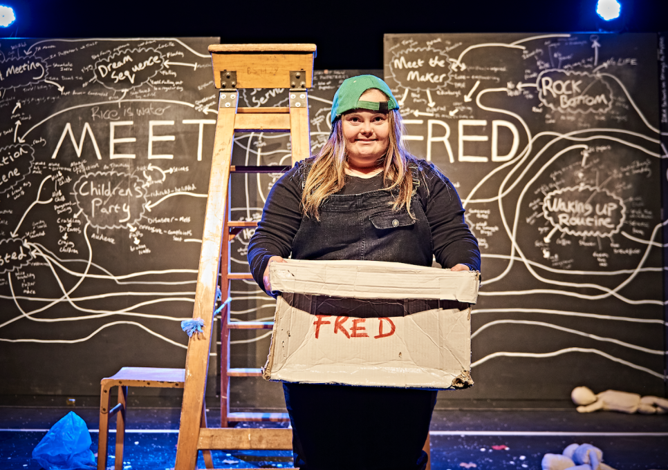 A woman in a green baseball cap is stood smiling, holding a box, in front of a blackboard with chalk diagrams and the writing 'MEET FRED'