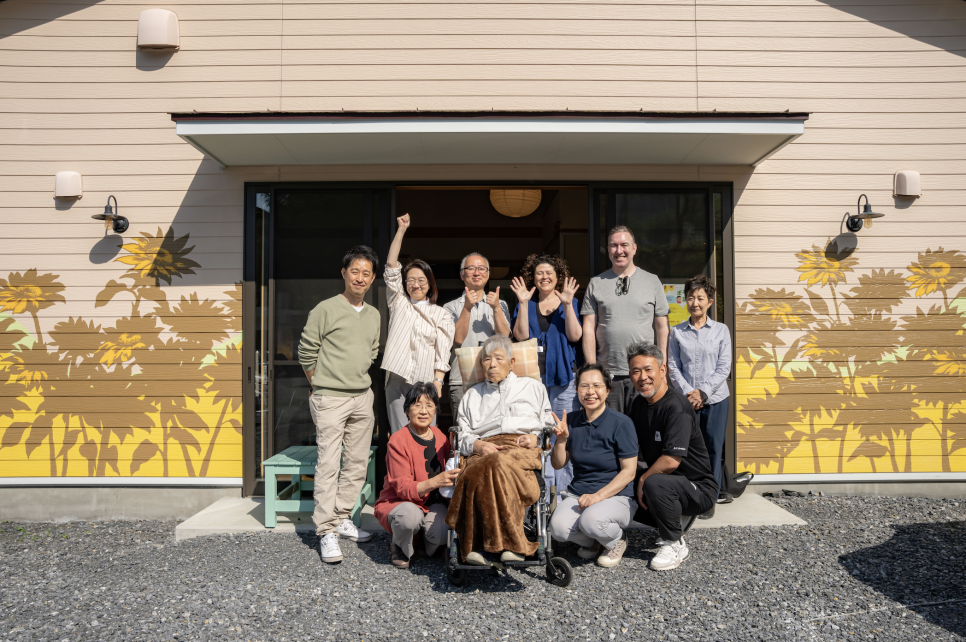 A group of people, an elderly man in a wheelchair at the front, smiling and waving in front of a building with sunflowers painted on it