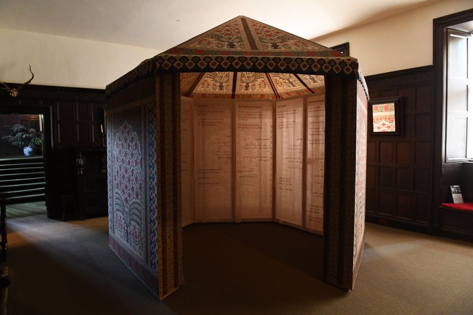 A free standing structure in the centre of a room panelled with dark wood. There is a missing panel in the structure to create an entrance. The walls and the roof of the structure are made from block printed fabric.