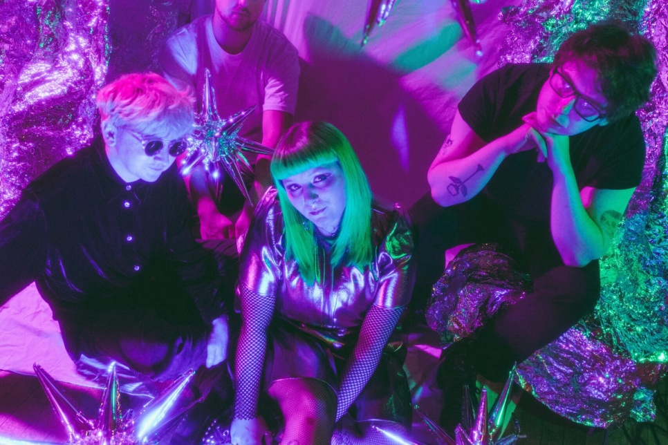 our people sit in a colourful, futuristic setting decorated with shiny metallic fabrics and star-shaped balloons. The scene is lit with bright pink, purple, and green lights. The person in front has green hair and wears a metallic outfit, while the others wear darker clothing and sunglasses or glasses, creating a vibrant, space-themed atmosphere.