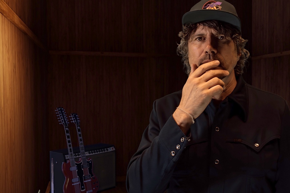 Male singer Gruff Rhys wearing a baseball cap and his hand over his mouth.