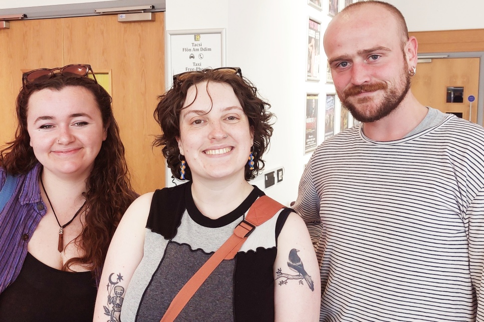 Colourful graphic: Two women and a man in their 20s pose for a photo in a theatre foyer, all three have pale skin. The two women have their hair pushed back by sunglasses on their heads, the man has a neat beard and striped tshirt / Graffig lliwgar. Dwy fenyw a dyn yn eu 20au yn sefyll am lun mewn cyntedd theatr, mae gan y tri groen golau. Mae gwallt y ddwy fenyw wedi'i wthio yn ôl gan sbectol haul ar eu pennau, mae gan y dyn farf daclus a chrys-t streipiog