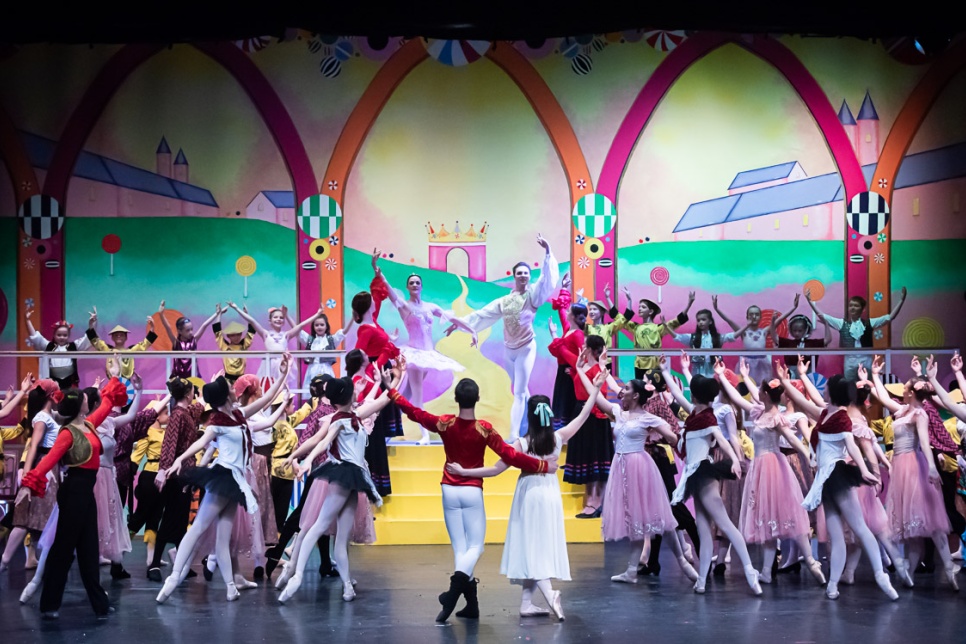 A large group of ballet dancers in colourful costumes onstage in front of a brightly painted set
