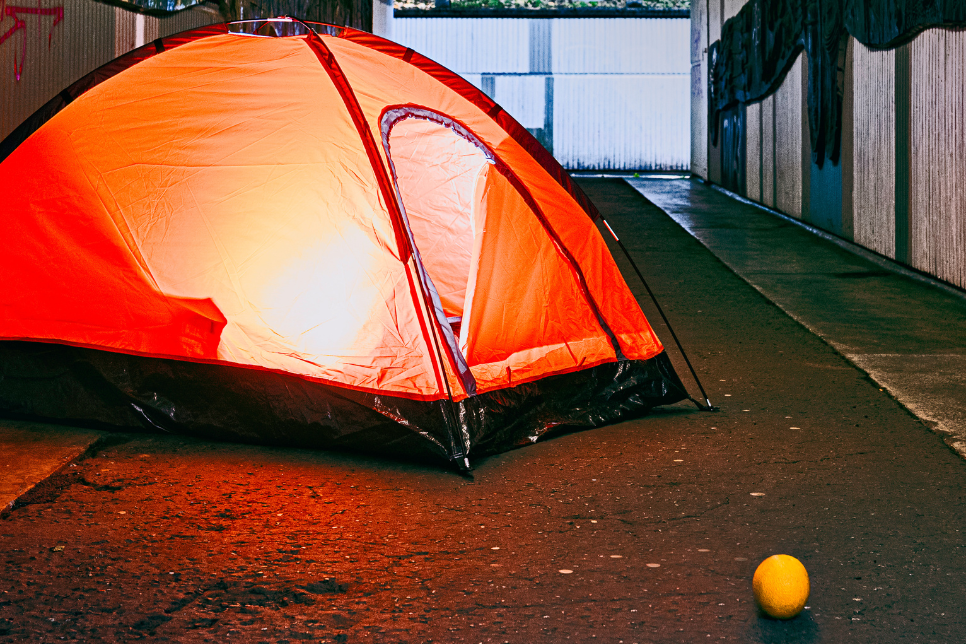 An orange pop up tent in a dark alley in the city. The tent is bright and lit from within.