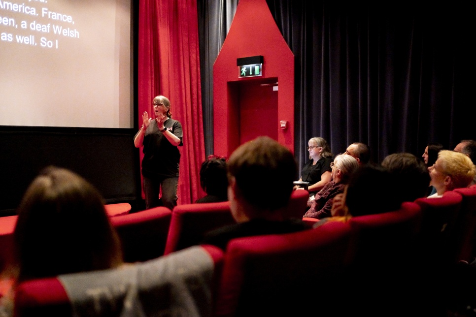 A woman dressed in black is stood in front of a cinema screen, signing in BSL to the audience.