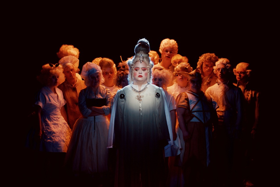 Still from Inside My Heart; a group of people stand together against a dark background in white clothing and Georgian style wigs, in orange lighting.