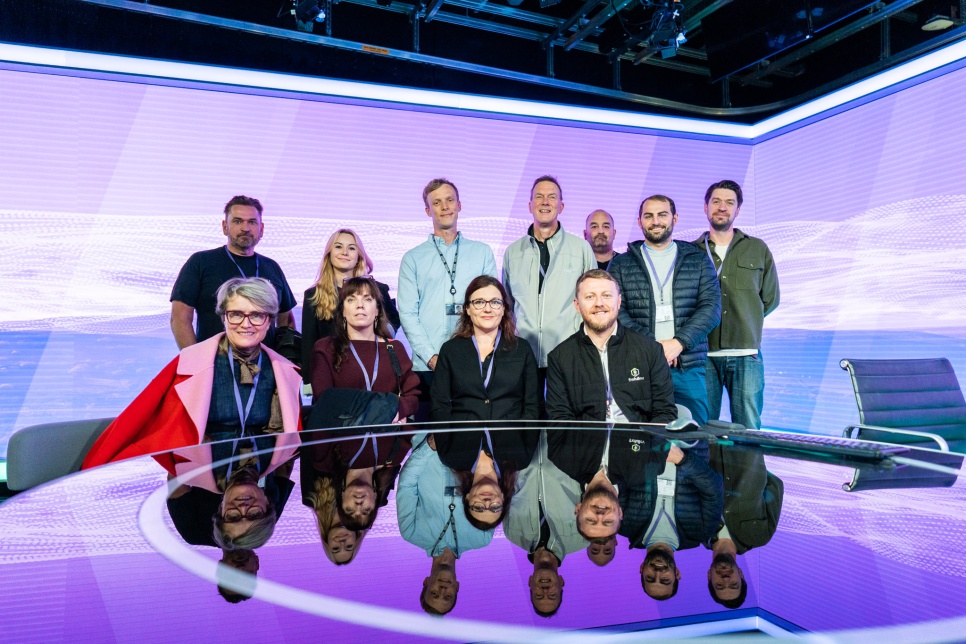 11 people sitting and standing around a reflective table in a TV news studio. 