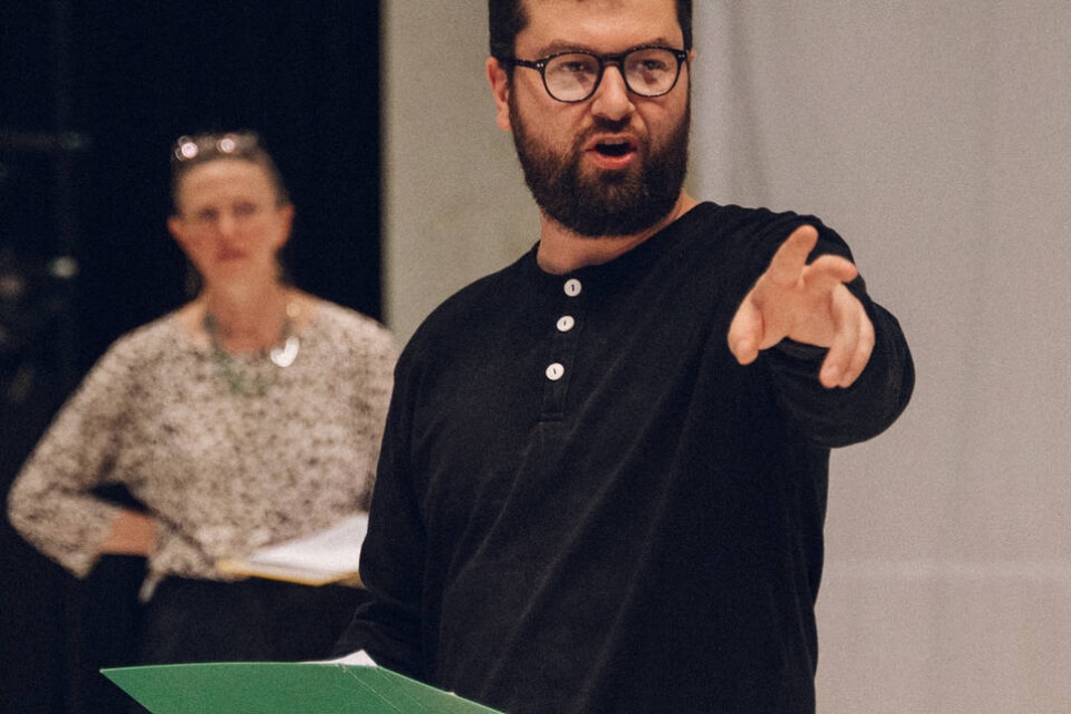 A picture of a man speaking. He is holding one hand in the air and holding a folder in the other. There is a woman standing in the background who is also holding a folder / Llun o ddyn yn siarad. Mae'n dal un law i fyny ac yn dal ffolder yn y llaw arall. Mae menyw yn sefyll yn y cefndir sydd hefyd yn dal ffolder.