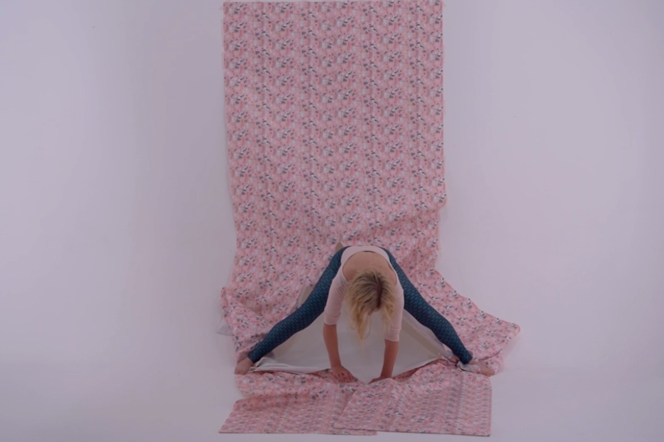 Artistic photograph involving a person, a white wall and floor, and a long patterned pink strip of material / Ffotograff artistig sy'n cynnwys person, wal a llawr gwyn, a stribed hir o ddefnydd pinc sydd â phatrwm