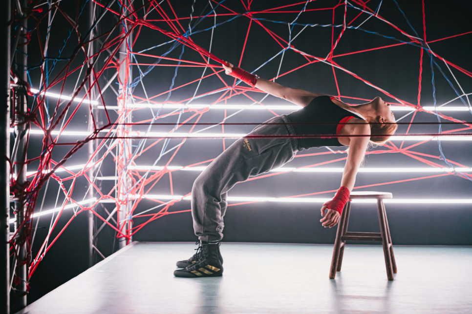 A platform with a person leaning backwards at an angle holding on to a part of a spiderweb of rope that raises up and over the platform. There is a stool placed behind the person / Llwyfan gyda pherson yn pwyso nôl ar ongl yn dal rhan o we-pry-cop o raff sydd yn codi lan a dros y llwyfan. Mae stôl wedi cael ei gosod tu ôl i’r person